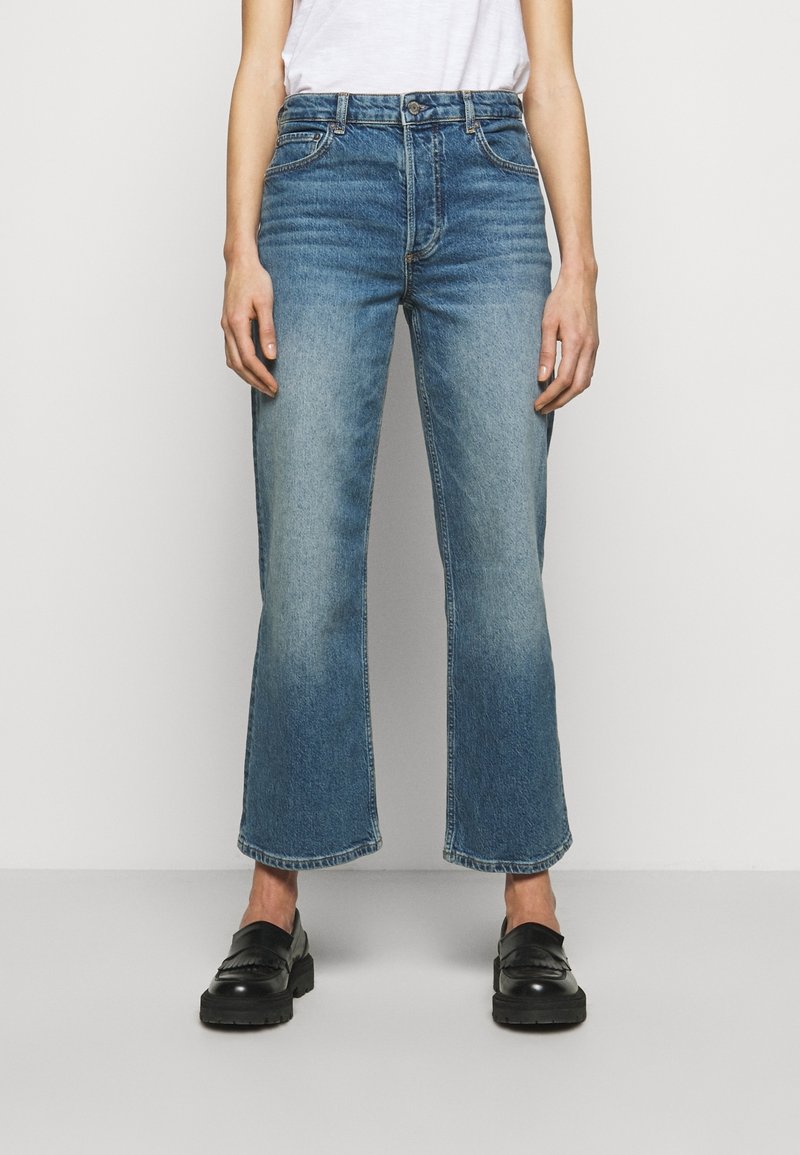 Personne portant un jean flare court taille haute délavé bleu, une chemise blanche rentrée et des mocassins noirs, debout devant un fond uni.