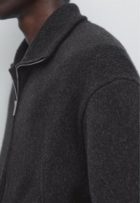 Zwarte zip-up cardigan van een gestructureerde wolmix. Heeft een geribde kraag en een gladde mouwstof, die een close-up zijaanzicht toont.