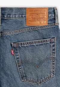 Poche arrière d'un jean en denim bleu avec des coutures orange, une étiquette rouge Levi's, et une étiquette en cuir marron Levi Strauss & Co. au-dessus de la poche.