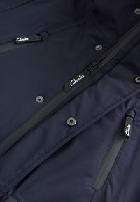 Veste bleu marine avec plusieurs fermetures éclair noires résistantes à l'eau et des boutons-pression en métal, avec le logo Clarks sur les tirettes.