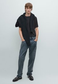 Sort crew-neck t-shirt, grå løst siddende jeans og sorte lædersko. Struktureret sort sweater draperet over skuldrene. Ensfarvet baggrund.