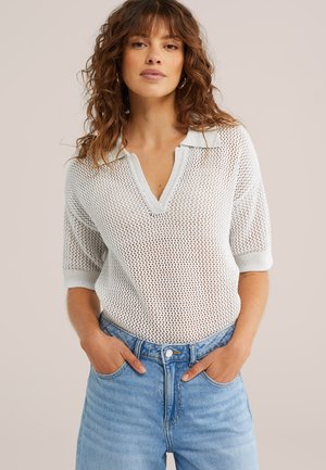 Mujer joven con cabello ondulado, vestida con una camiseta blanca de punto de manga corta y vaqueros azul claro, con las manos en los bolsillos, fondo neutral.