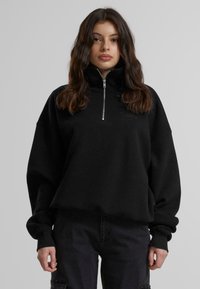 Svart hettegenser med glidelås laget av myk stoff, med høy krage, senkede skuldre og subtilt logotekst på fremsiden.