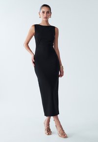 Black midi dress with a fitted silhouette, sleeveless design, and boat neckline. Paired with gold strappy sandals, against a light backdrop.