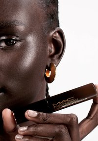 Brun concealer tube holdt mot mørk hud, med gulltekst og en blank finish; modellen har gull hoopsmykker. Jevn, glatt tekstur.