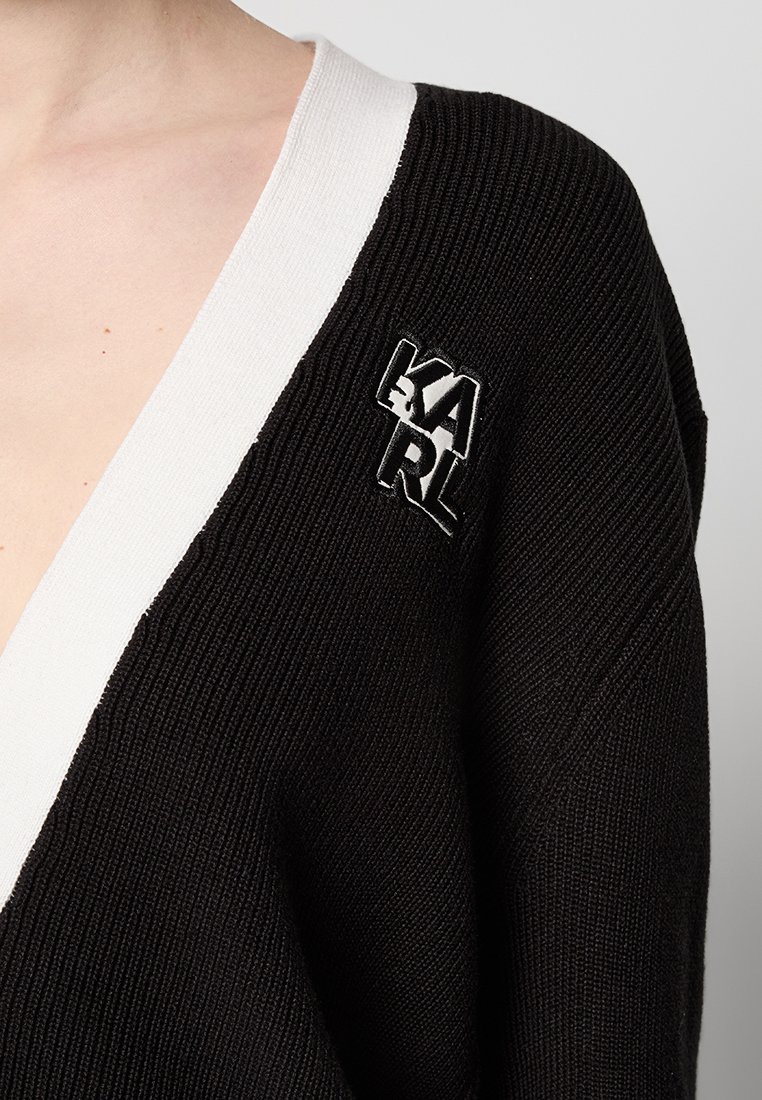 Cardigan noir en tricot avec un décolleté en V profond, une bordure blanche, et un logo « KARL » brodé en noir et blanc situé sur le côté gauche.