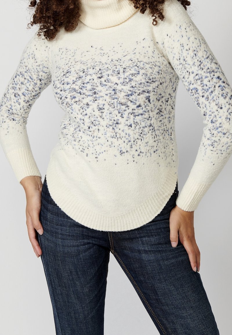 Maglione a collo alto color crema con un motivo testurizzato, caratterizzato da macchie blu. Tessuto morbido e lavorato a maglia con orlo curvo. Abbinato a jeans scuri.