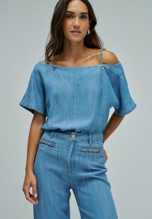 Mujer con una blusa azul claro sin hombros con tirantes de cadena y vaqueros de talle alto a juego con detalles decorativos en los bolsillos.
