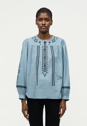 Kobieta zwrócona przodem, ubrana w jasno-niebieską haftowaną bluzkę z długimi bufkami i czarne spodnie na jednolitym tle.