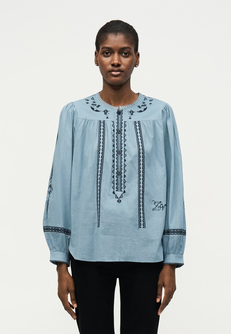 Γυναίκα που κοιτάζει μπροστά, φοράει ανοιχτό μπλε κεντημένο μπλουζάκι με μακριά φουσκωτά μανίκια και μαύρο παντελόνι σε απλό φόντο.