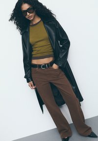 Zwarte leren trenchcoat, olijfgroene fitted top met bruine afwerking, en bruine wijde broek. Accessoires zijn onder andere een zwarte riem en grote zonnebril.