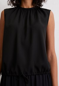 Vrouw in een mouwloze zwarte blouse met een geplooide halslijn en een trekkoord onderaan, getoond van nek tot taille tegen een neutrale achtergrond.