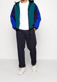 Color-blocked jacket with green, blue, and purple panels. Features a front zipper, drawstring hood, and elastic cuffs. Paired with navy pants and white sneakers.