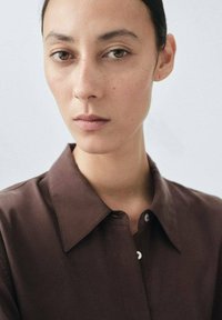 Camicia marrone con colletto, caratterizzata da una texture liscia e dettagli con bottoni; modello con capelli scuri e lisci, sfondo neutro e trucco naturale.
