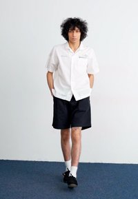 White short-sleeved button-up shirt with embroidered detailing, paired with black shorts, white socks, and black shoes, standing on a blue carpet.