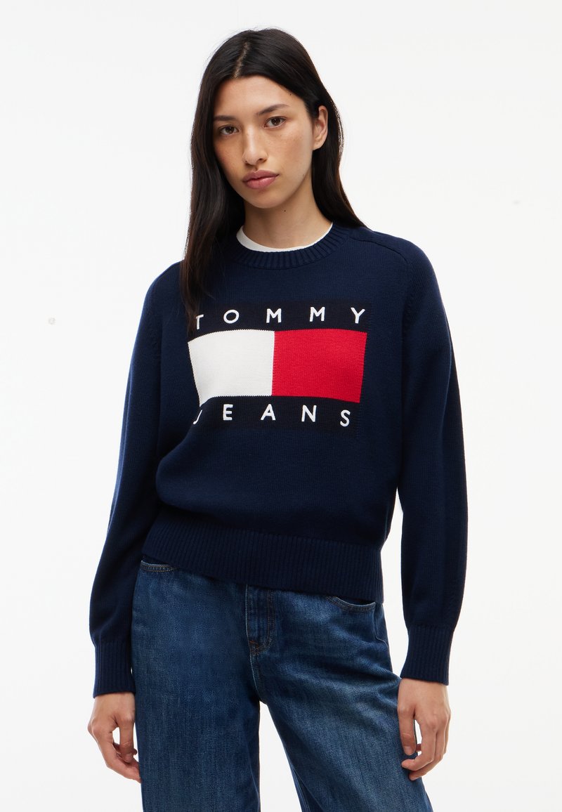 Jeune femme aux longs cheveux foncés portant un pull bleu marine Tommy Jeans et un jean bleu sur un fond blanc uni.