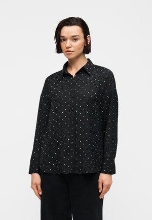 Jeune femme aux cheveux courts et foncés portant une chemise noire à manches longues avec de petits pois blancs, debout devant un fond uni.