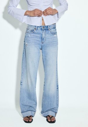 Person iført lyseblå vidde jeans med mindre slid, en stribet skjorte med knapper og sorte klipklappere mod en ensfarvet baggrund.