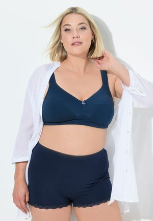Reggiseno blu navy con spalline larghe e bordi in pizzo, con un accento floreale centrale. Abbinato a slip boyshorts blu navy. Camicia leggera bianca.