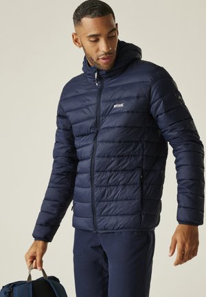 Mężczyzna w granatowej kurtce puchowej i spodniach, trzymający niebieski plecak za rączkę, patrzący w dół w lewo.