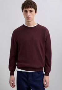 Suéter de punto color burdeos con mangas largas y cuello redondo, combinado con una camiseta blanca. Presenta un detalle sutil de logotipo en el pecho.