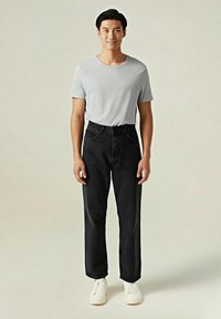 Joven de pie, vistiendo una camisa de manga corta gris claro, pantalones negros de talle alto y zapatillas blancas frente a un fondo beige liso.