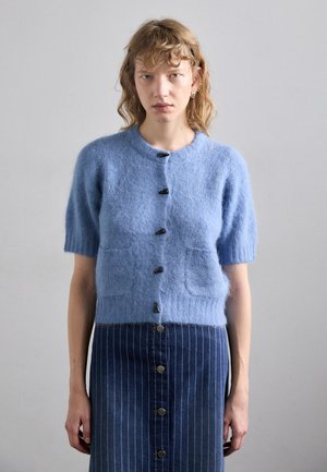 Cardigan bleu clair à manches courtes en matière pelucheuse avec cinq boutons foncés, présentant deux poches avant. Assorti à une jupe en jean foncé.
