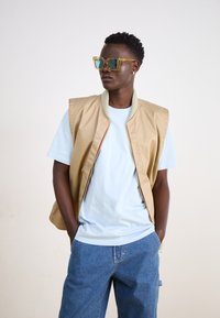 Beige ujjatlan mellény egy világoskék póló felett, kék farmer sorttal párosítva. A modellen túlságosan nagy sárga napszemüveg van, és texturált frizurával rendelkezik.