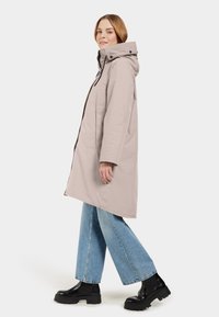 Cappotto impermeabile beige chiaro con cappuccio, tasche frontali e zip. Abbinato a jeans blu larghi e stivali platform neri.