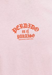 Gaiši rozā kokvilnas t-krekls ar drosmīgu, oranžu tekstu, kas lasa "PERDIDO en el PARAÍSO", ar "BIENVENIDOS A PUERTO RICO" mazākā fontā zem tā.