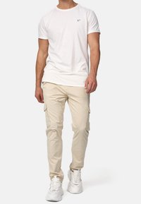 Valkoinen puuvillapaita, jossa on pieni logo rinnassa, yhdistettynä beigekiin kargo-housuihin, joissa on sivutaskut. Mallilla on päällään valkoiset lenkkarit.