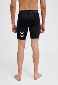 Čovjek stoji bos, nosi crne sportske kompresijske kratke hlače s bijelim chevron logotipima na lijevoj nozi, prikazan s leđa.