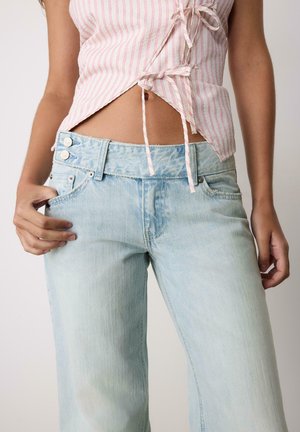 Donna che indossa jeans a vita alta di colore azzurro chiaro con gamba larga e un top senza maniche a righe rosa e bianche con laccetti sul davanti, su sfondo semplice.