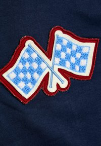 Țesătură bleumarin cu un patch decorativ brodat reprezentând două steaguri în carouri albastre încrucișate, înconjurate de o cusătură roșie.