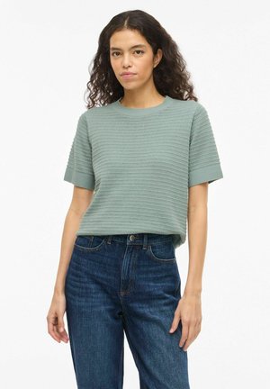 Kvinde med krøllet mørkt hår iført en tekstureret salviegrøn kortærmet sweater og højtaljede blå jeans, stående foran en hvid baggrund.