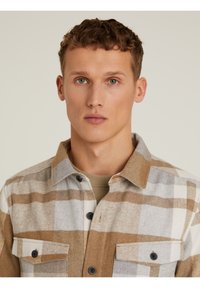 Camisa de cuadros en tonos beige, marrón y gris, con un frente abotonado, dos bolsillos en el pecho y un cuello clásico. Textura suave.