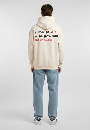 Felpa crema con un grande cappuccio e polsini a coste, con testo giocoso in nero e rosso sulla schiena. Abbinata a jeans azzurri chiari e sneakers nere.