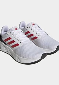 Chaussures de sport blanches avec une tige en mesh, design à trois bandes rouges, col rembourré et semelle extérieure en caoutchouc noir. Légères avec un système de laçage.