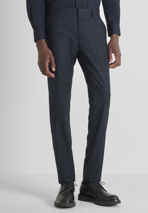 Hombre vestido con pantalones de vestir oscuros de cuadros ajustados y zapatos de cuero negro con cordones, de pie contra un fondo claro y liso.