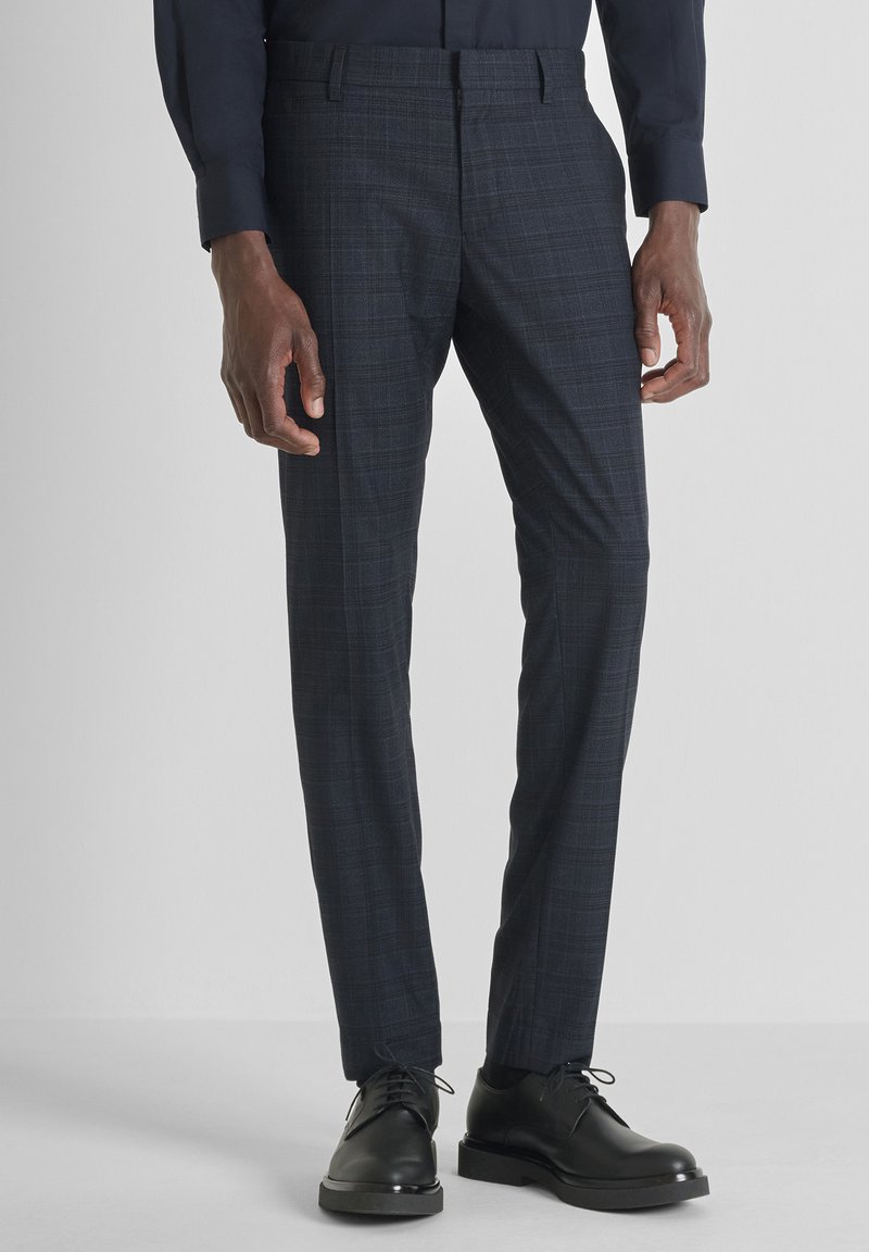 Muškarac u uskim tamnim kariranim odijelima i crnim kožnim cipelama s vezicama, stoji naslonjen na svijetlu ravnu pozadinu.