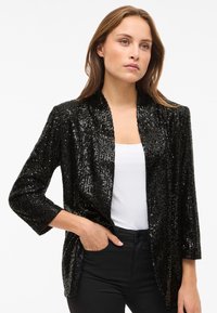 Zwarte blazer met pailletten, een sjaalkraag, driekwart mouwen, een open voorkant en een getextureerd oppervlak. Gecombineerd met een wit topje en zwarte broek.