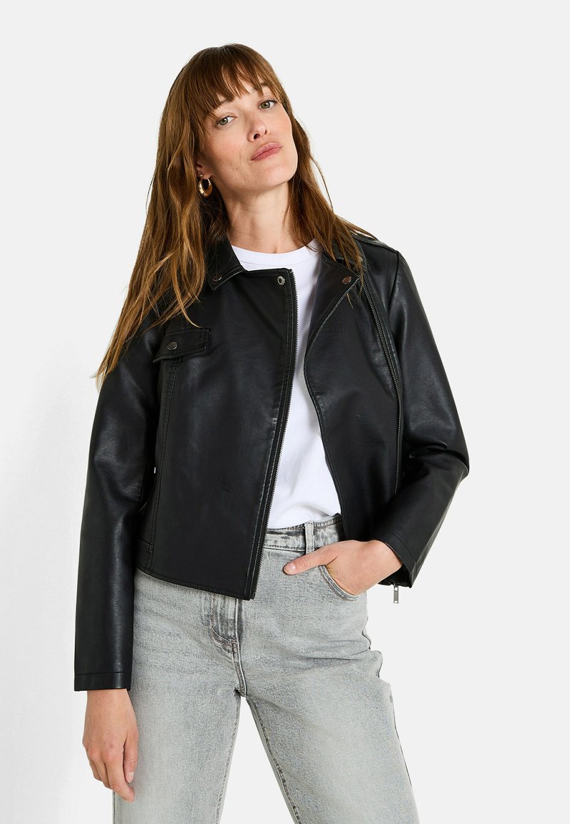 Femme avec de longs cheveux bruns portant des boucles d'oreilles en or, une veste en cuir noire sur une chemise blanche, et un jean gris clair avec une main dans la poche.