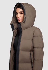 Brun puffjacka med hög huva, med svarta detaljer och dragkedjor. Slät yta med kviltad textur för isolering.