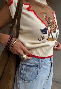 Άτομο που φοράει πουλόβερ χωρίς μανίκια με σχέδιο καουμπόη και άλογο, ανοιχτό μπλε τζιν με στρογγυλεμένες τσέπες και πολλούς δαχτυλίδια και βραχιόλια.