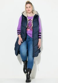 Marinblå pufferväst med huva och fluffig foder, lagd över en lila grafisk sweatshirt med randiga ärmar, i kombination med blå jeans och svarta stövlar.