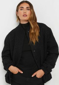 Veste bomber noire en textile texturé avec un col arrondi, fermeture à boutons-pression, et ourlet côtelé. Portée par-dessus un haut noir, les mains dans les poches.