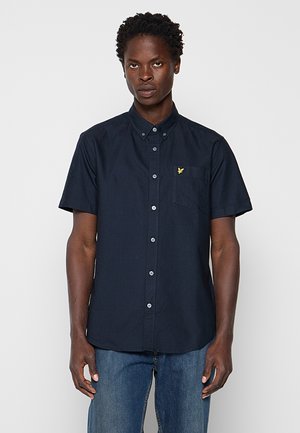 Mann trägt ein marineblaues kurzärmliges Hemd mit gelbem Vogel-Logo auf der Brusttasche und blaue Jeans, steht frontal.
