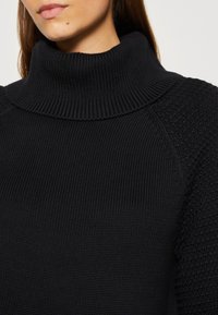 Pull noir en maille avec un col montant côtelé. Tissu texturé avec un motif sur les manches et les coutures des épaules. Design épais et confortable.