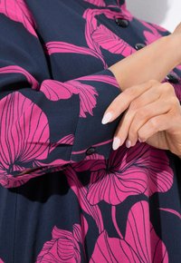 Navyfarbendes Hemd aus Stoff mit großem pinkfarbenem Blumenmuster, mit Knopf am Ärmel und glatter Textur. Nahaufnahme von Arm- und Handpositionierung.