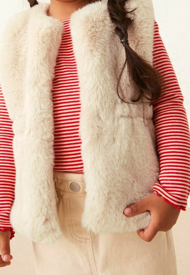 Gilet beige peloso indossato sopra una maglietta a maniche lunghe rossa e bianca a righe, abbinato a pantaloni beige. Il gilet presenta una texture morbida e pelosa.